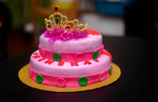Two-tier fondant cake design with a tiara on top, decorated with icing flowers. Delicious pink-red color-themed round cake on yellow cake tray against the dark background.