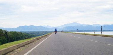 Düz bir yol, uzaklıktaki sisli dağ sırasına, Udawalawa barajının tepesine inşa edilen yola, soldaki ormana ve barajın sağındaki Udawalawa rezervuarına çıkar..