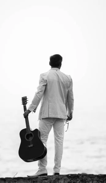 White dressed man holding a guitar in one hand, view from the back in the beach, black and white photograph.