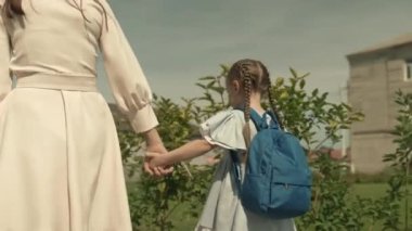 Anne, küçük kızını sırt çantasıyla okula götürüyor, çocuğa anaokuluna kadar eşlik ediyor, okul bahçesinde yürüyor, birinci sınıf öğrencisi, mutlu bir aile, okul çantalı bir çocuk ve annesi.