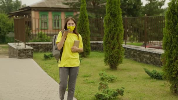 Ein junges Mädchen geht über den Schulhof und telefoniert mit einem Lehrbuch in der Hand, ein Schulmädchen mit einem Rucksack kommuniziert online auf einem Smartphone, ein moderner SchulmädchenTeenager — Stockvideo