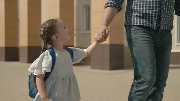 Vader houdt gelukkige dochter bij de hand, begeleiden klein kind met rugzak naar school, studeren in kleuterschool voorbereiding, lopen door schoolplein, eerste klas student met schooltas op zijn schouders — Stockfoto