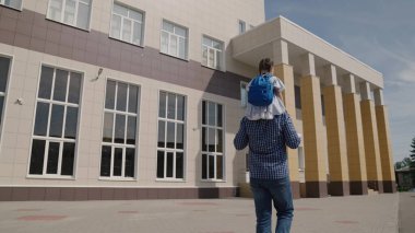 Baba omuzlarında sırt çantalı küçük bir çocuk, okul çantalı bir kız okul bahçesinde babasına biniyor, mutlu bir aile bir ilkokul, birinci sınıf öğrencisi sınıfa gidiyor.
