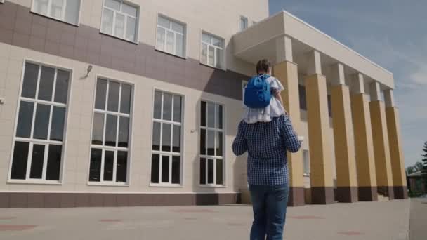 Papai carrega em seus ombros criança pequena com mochila para a escola, menina com bolsa de escola monta seu pai ao redor do pátio da escola, família feliz uma escola primária, primeiro graduador com os pais vai para a aula — Vídeo de Stock