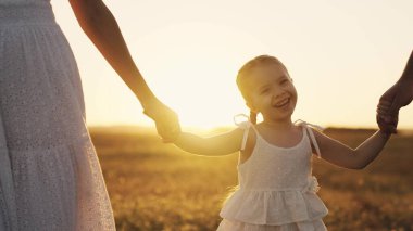 Mutlu bebek gün batımında anne, baba ve çocukla birlikte gülümsüyor. Güneşte yürüyorlar. Çocuklarını elleriyle tutuyorlar, küçük kızlarına bakıyorlar.