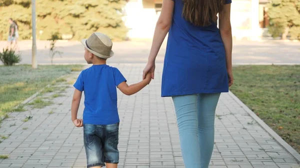 Mutter führt ihren kleinen Sohn an der Hand durch Stadtpark, glückliche Familie, Ruhetag mit Kind, Sommerreise mit Kind, Hand in Hand halten, aufeinander achten, Junge hat Spaß, Kindheitstraum — Stockfoto