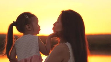 Küçük kız gün batımında annesini öpüyor, mutlu aile, çocukluk hayali, anne sevgili kızına güneş ışığında sarılıyor, çocuklar hafta sonları ebeveynleriyle takılıyor, bebeği dudaklarından öpüyor.