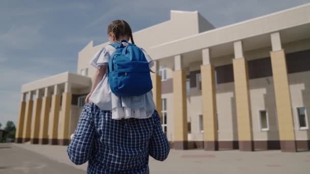Dad carries on his shoulders little kid with backpack to school, girl with school bag rides her father around the schoolyard, happy family an elementary school, first grader with parent goes to class — Stock Video