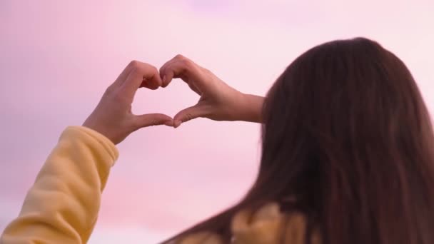 Chica muestra la forma del corazón al atardecer rosa en el cielo, San Valentín romántico, amanecer mañana de nubes rosadas, niña sueña con el amor, felices vacaciones de verano, día festivo libre en la naturaleza. — Vídeos de Stock