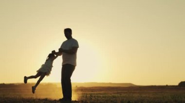 Baba, gün batımında küçük bir kızı gökyüzünde daire çiziyor. Neşeli bir çocuk güneş ışınlarıyla dünyanın üzerinde süzülüyor. Babasıyla çocuk pilotçuluk oynuyor, mutlu aile, kız ve erkek silueti.