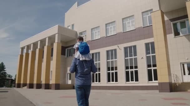 A little kid rides on his fathers shoulders to school with a backpack, a girl with a school bag and dad goes to first grade, a happy family, getting primary education at school, a child with parent — Stock Video