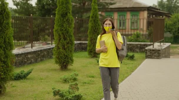 Młoda dziewczyna przechodzi przez szkolny dziedziniec i rozmawia przez telefon trzymając podręcznik w rękach, uczennica z plecakiem komunikuje się online na smartfonie, nowoczesna nastolatka uczennica — Wideo stockowe