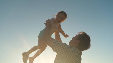 Mutlu baba güneşin parıltısıyla bir çocuğu gökyüzüne fırlatır, çocuk bir uçak gibi yükseğe uçar ve güler, küçük kız spa ile açık havada oynar, mutlu aile kavramı