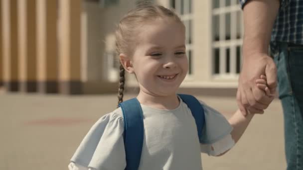 Papá está llevando a una pequeña hija a la escuela, una chica con una mochila está sosteniendo la mano de sus padres, un sistema de educación escolar para niños, una familia feliz, un niño alegre con un padre camina a través de la — Vídeos de Stock