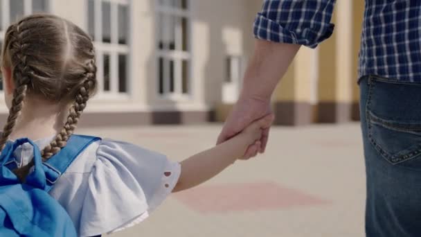 Père tient sa fille heureuse par la main, accompagne le petit enfant avec un sac à dos à l'école, étudie la préparation préscolaire, traverse la cour d'école, élève de première année avec un sac d'école sur les épaules — Video