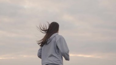 Spor giyimli bir kız gün batımında gökyüzüne doğru koşar, kadınlar uzun mesafe koşar, sağlıklı yaşam tarzı, zayıf bir vücut ve sağlıklı bir kalp için kardiyo eğitimi, ilk olmak için çabalar, şafakta koşar.