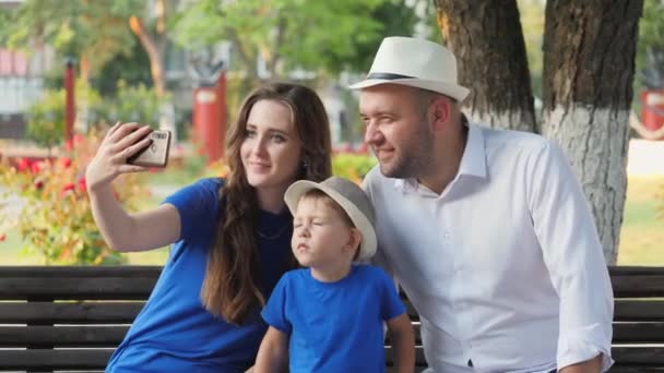 Mutlu bir aile gülümsüyor telefonda selfie çekiyor, baba ve çocuk akıllı bir telefonla fotoğraf çekiliyor, anne, baba ve oğul internet üzerinden iletişim kuruyor, küçük çocuğu ve şehir insanlarını seviyor. — Stok video