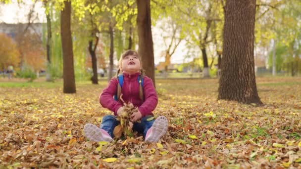 Dívka s batohem hází suché podzimní listí přes hlavu, školní taška na ramenou školačky, dítě v městském batohu hraje se žlutým listím, šťastná malá dětská příroda venku — Stock video
