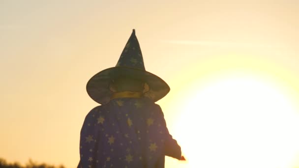 Menino em um chapéu evoca varinha mágica ao pôr do sol, alucinar, acreditar em crianças mágicas, criança brinca no brilho do sol, fantasia de carnaval infantil para férias, uma roupa festiva astranom, sonho de infância — Vídeo de Stock