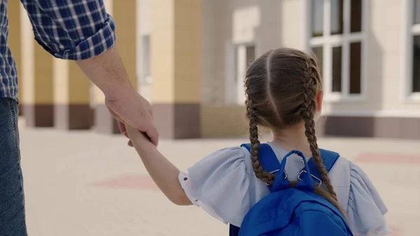 Otec drží šťastnou dceru za ruku, doprovází malé dítě s batohem do školy, studuje v předškolní přípravě, chodí po školním dvoře, student první třídy se školní brašnou na ramenou — Stock fotografie