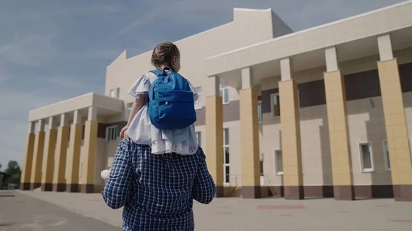 Papai carrega em seus ombros criança pequena com mochila para a escola, menina com bolsa de escola monta seu pai ao redor do pátio da escola, família feliz uma escola primária, primeiro graduador com os pais vai para a aula — Fotografia de Stock