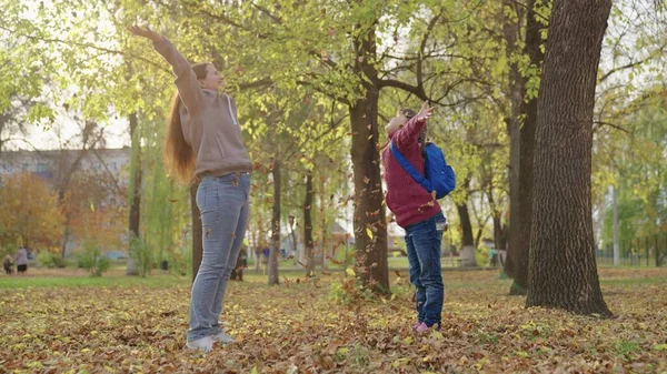 Kızı ve annesi sonbahar sarısı yaprakları fırlatıp gülüyorlar, okul çantalı çocuk yapraklarla oynuyor, mutlu aile, çocukluk hayalleri, birinci sınıf liseli kız ve ebeveyni neşeli.