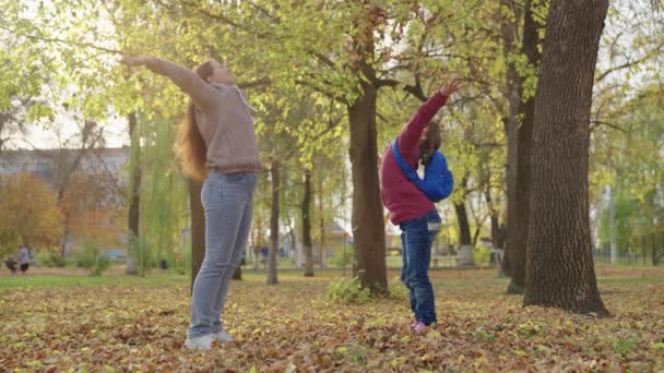 Дочь и ее мать бросают осенью желтые листья вверх и смеяться, ребенок со школьной сумкой играет с осенью листья, жизнь счастливая семья, детские фантазии мечты, первоклассная школьница с родителями весело — стоковое видео