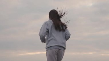 Spor giyimli bir kız gün batımında gökyüzüne doğru koşar, kadınlar uzun mesafe koşar, sağlıklı yaşam tarzı, zayıf bir vücut ve sağlıklı bir kalp için kardiyo eğitimi, ilk olmak için çabalar, şafakta koşar.