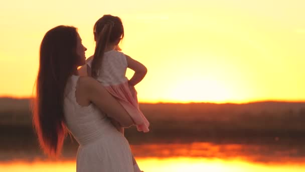 Mam en meisje tonen de zon in de lucht en glimlach, gelukkig kinderleven, kind met moeder aan de kust bij zonsondergang, houd klein kind in haar armen, baby heeft plezier aan de rivier in de open lucht van de natuur van zonnen stralen. — Stockvideo