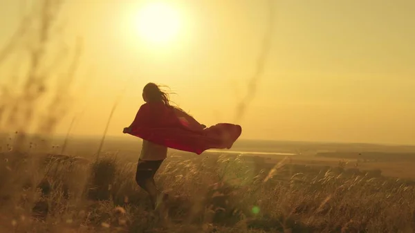Happy teenage girl runs playing superhero at sunset, child on a green field in a red raincoat toy in the rays of light, childrens dream game, kid dreams of becoming a superhero in glare of the sun — Stock Photo, Image