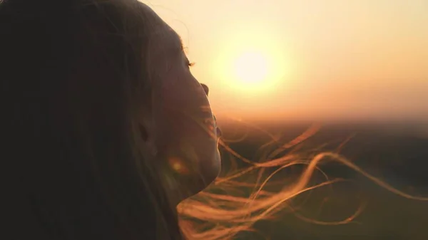 Young girl looks at sunset and prays, religious person, her hair is flying in wind in glare of the sun, to believe in goodness, a womans dream of love, to think and ask for forgiveness from heaven — Stock Photo, Image