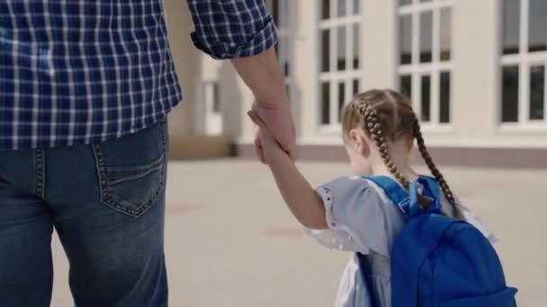 Vater hält glückliche Tochter an der Hand, begleitet kleines Kind mit Rucksack zur Schule, lernt in Vorschulvorbereitung, geht über Schulhof, Erstklässler mit Schultasche auf den Schultern — Stockvideo