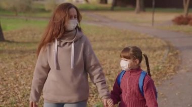 Sırt çantalı küçük bir çocuk okul bahçesinde tıbbi maskeler takıyor, anne çocuğu ders için neşeli bir şekilde okula koşuyor, çocukluğu koronavirüs salgınından korunuyor, covid 19, birinci sınıf.