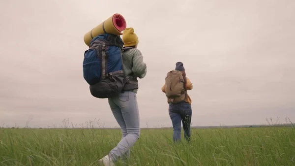 Chicas hipster activas felices con mochilas en la espalda viajan a través de un hermoso valle, una mujer milenaria en un campo verde, excursiones, senderismo en el sendero, recreación al aire libre —  Fotos de Stock