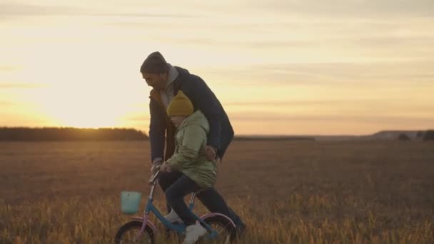 Bambino impara ad andare in bicicletta con il padre al tramonto, famiglia felice, sogno d'infanzia di imparare ad andare in bicicletta, pedali bambino al sole, ragazza e papà ridere divertente durante il gioco, giorno dei padri — Video Stock