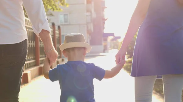 A little boy holding his mother and fathers hands in the rays of the sun, walking around the city with the child, a happy family, mom, dad and kid in glare of the sun, childhood dream of babys life — Stock Photo, Image