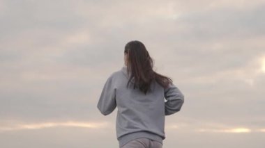 Spor giyimli bir kız gün batımında gökyüzüne doğru koşar, kadınlar uzun mesafe koşar, sağlıklı yaşam tarzı, zayıf bir vücut ve sağlıklı bir kalp için kardiyo eğitimi, ilk olmak için çabalar, şafakta koşar.