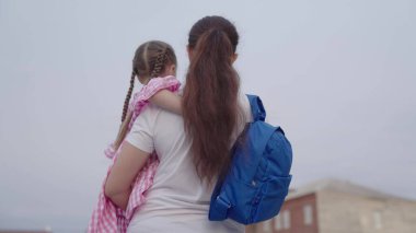 Anne kucağında sırt çantasıyla küçük bir kızı okula taşıyor, ilkokulda bir çocuğa öğretmenlik yapıyor, mutlu bir aile, okul çantasıyla bir çocuğu uğurluyor, gelecekle ilgili bilgi topluyor.