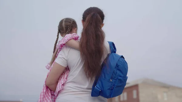 Mãe carrega uma filhinha com uma mochila nos braços para a escola, ensinando uma criança na escola primária, uma família feliz, vendo uma criança com uma bolsa de escola, ganhando conhecimento da vida futura — Fotografia de Stock