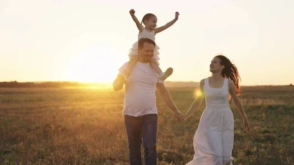 Happy family, mother, father and little child, kid sits on shoulders dad, childhood dream of flying, parents with their daughter run at sunset, adult man and woman laugh playing with baby in sunlight — Stock Photo, Image