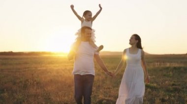 Mutlu aile, anne, baba ve küçük çocuk, çocuk omuzlarında oturur baba, çocukluk hayali uçmak, ebeveynler kızlarıyla gün batımında koşar, yetişkin erkek ve kadın gün ışığında bebekle oynarken güler.