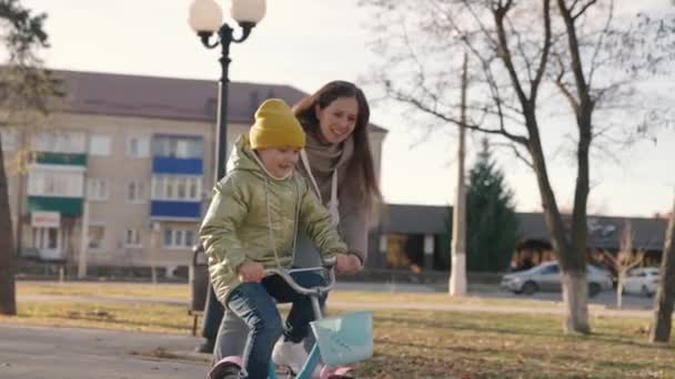 Mother teaches little child to ride bike in the city, kid rides on sidewalk, happy family, baby plays and laughs with mom, learn ride, make childhood dream come true girls, daughter and mom are funny — Stock Video