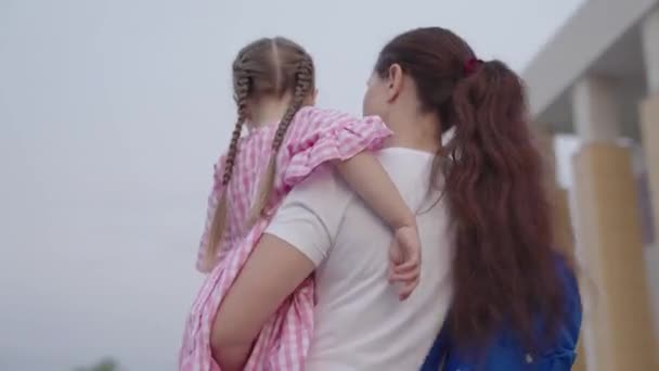 Madre lleva a una pequeña hija con una mochila en sus brazos a la escuela, enseñando a un niño en la escuela primaria, una familia feliz, despedir a un niño con una bolsa de la escuela, adquirir conocimiento de la vida futura — Vídeo de stock