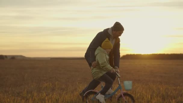 Отец учит маленького ребенка ездить на двухколесном велосипеде на солнце, счастливая семья, веселая детская жизнь, ребенок и папа вместе на выходных весело провести время на велосипеде, играть в игру с родителями на закате — стоковое видео