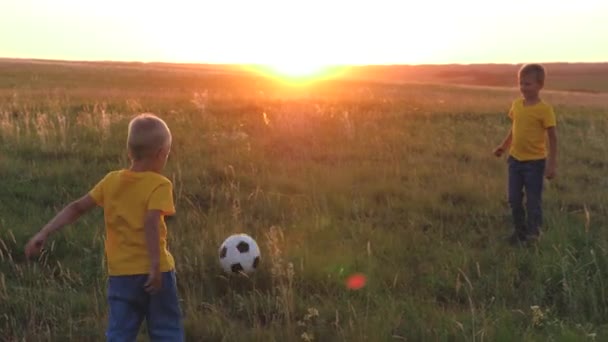 Meninos jogar futebol em um gramado verde ao pôr do sol, um campo de futebol, uma criança com uma bola ao sol, divirta-se com amigos em uma equipe, crianças felizes, sonho de infância de ser um jogador de futebol, filhos fantasia — Vídeo de Stock