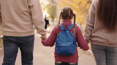 Anne ve baba küçük bir kızı sırtında sırt çantasıyla tutarlar, mutlu bir aile, sonbaharda okul bahçesinde yürürler, anne, baba ve çocuk omuzlarının üzerinde çanta taşırlar, birinci sınıftaki çocuğa eşlik ederler.