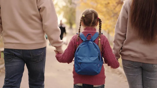 Mamma e papà tengono una piccola scolaretta con uno zaino per mano, famiglia felice, camminano attraverso il cortile della scuola in autunno, mamma, papà e bambino con borsa sulle spalle, accompagnano il bambino in prima elementare. — Foto Stock