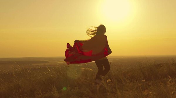 Happy teenage girl runs playing superhero at sunset, child on a green field in a red raincoat toy in the rays of light, childrens dream game, kid dreams of becoming a superhero in glare of the sun.