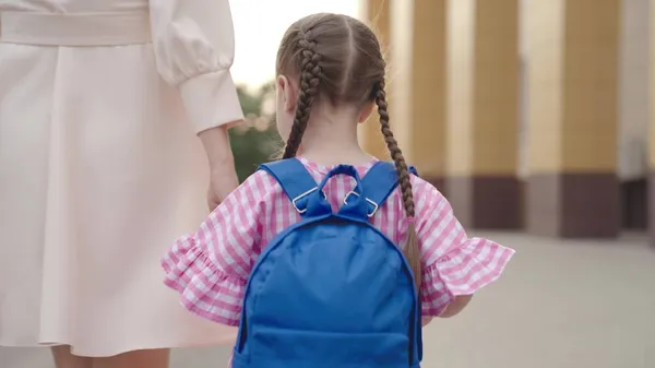 Uma criança pequena com uma mochila segura sua mãe pela mão e vai para a escola, a mãe leva uma criança com uma bolsa de escola ao redor do pátio da escola, família feliz, estudante da primeira série, educação pré-escolar primária — Fotografia de Stock