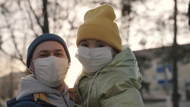 Face close-up in mask, little girl in arms of her dad in medical mask smiles in rays sunny city, child with her dad at sunset, to protect herself from coronavirus pandemic, covid 19, code is laughing — Stock Video